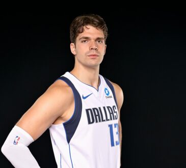 Dallas Mavericks forward Tarik Biberović stands with a hand on his hip in a white Dallas jersey against a black backdrop.