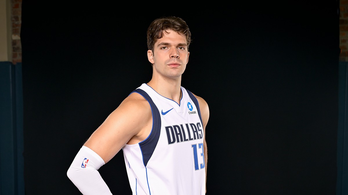 Dallas Mavericks forward Tarik Biberović stands with a hand on his hip in a white Dallas jersey against a black backdrop.