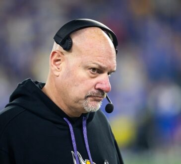 Vikings assistant coach Mike Pettine, wearing a headset and team hoodie, looks on from the sideline.