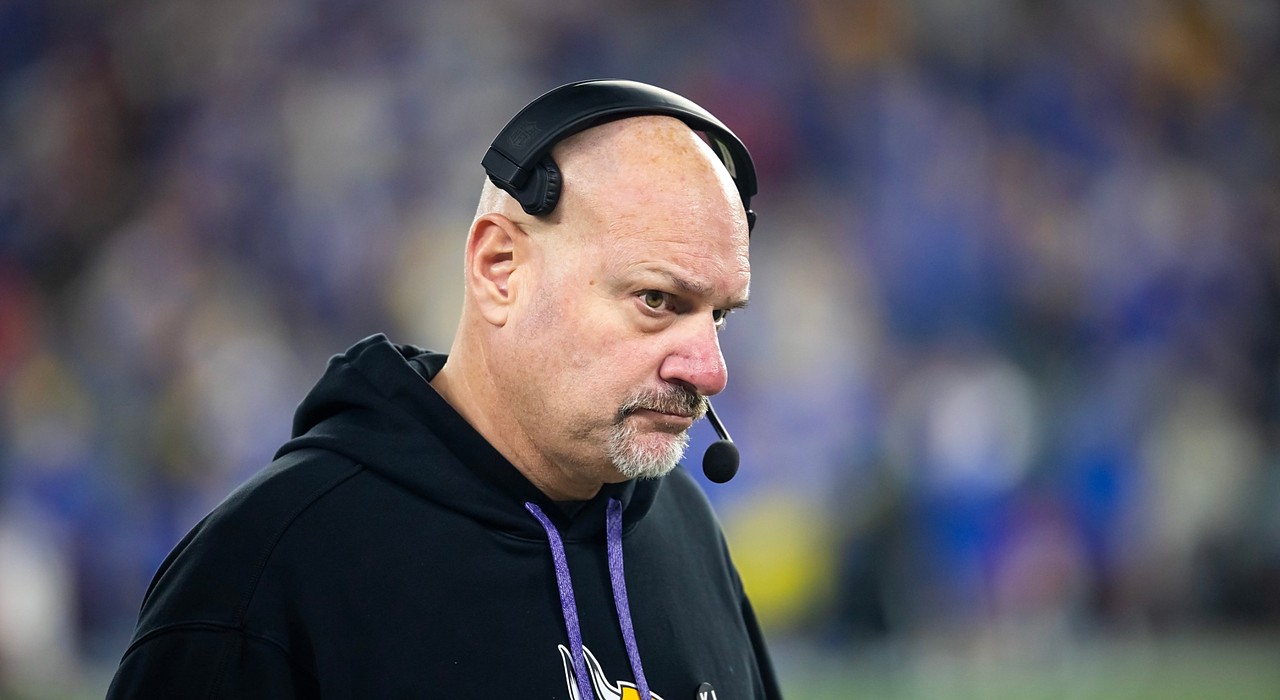 Vikings assistant coach Mike Pettine, wearing a headset and team hoodie, looks on from the sideline.