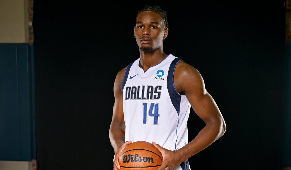 Mavericks forward Morez Johnson Jr. holds a basketball at his waist while posing in his white home jersey against a black backdrop.
