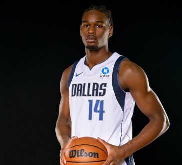 Mavericks forward Morez Johnson Jr. holds a basketball at his waist while posing in his white home jersey against a black backdrop.