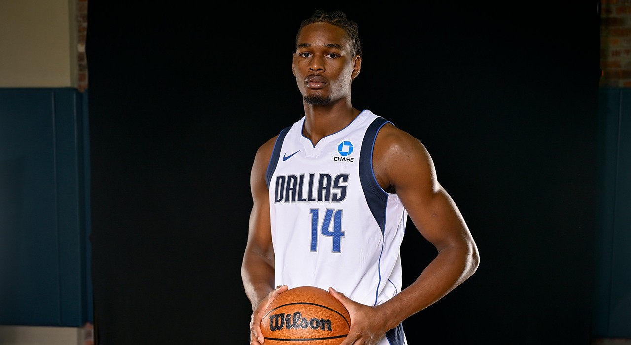 Mavericks forward Morez Johnson Jr. holds a basketball at his waist while posing in his white home jersey against a black backdrop.