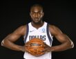 Dallas Mavericks center Moussa Cissé grips a basketball at his chest in a white Dallas jersey against a black backdrop.