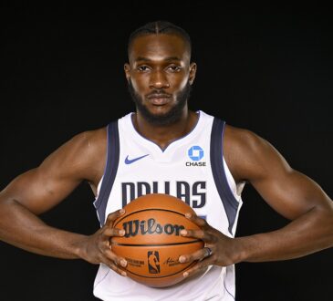 Dallas Mavericks center Moussa Cissé grips a basketball at his chest in a white Dallas jersey against a black backdrop.