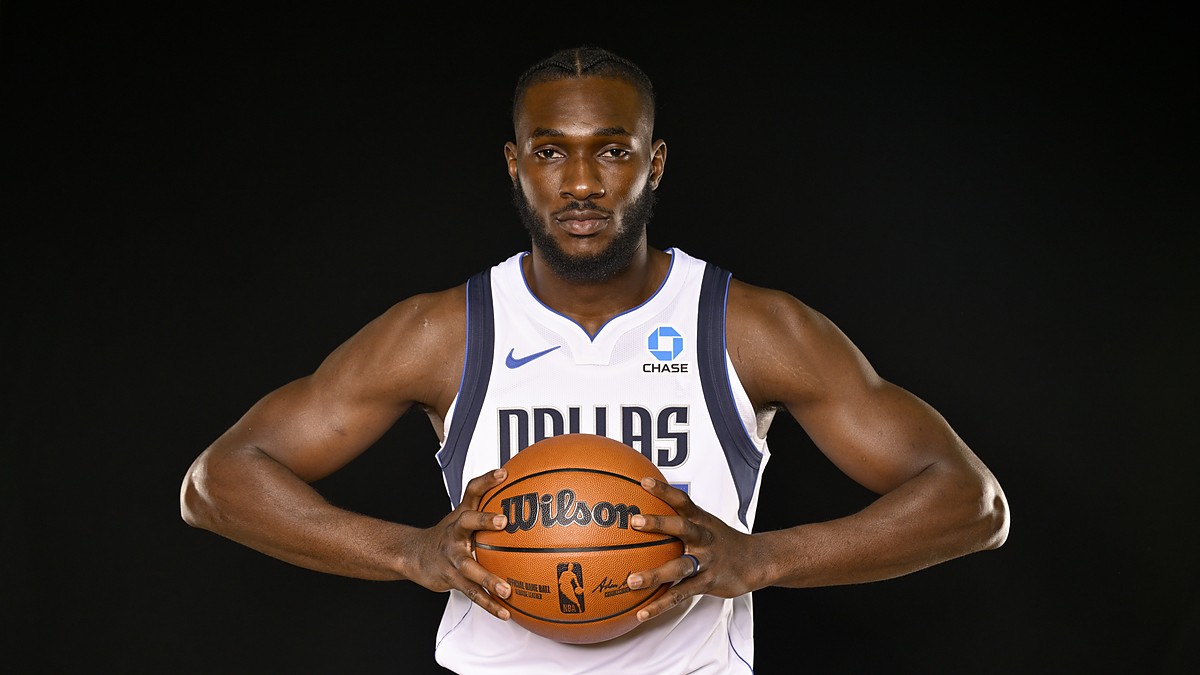 Dallas Mavericks center Moussa Cissé grips a basketball at his chest in a white Dallas jersey against a black backdrop.