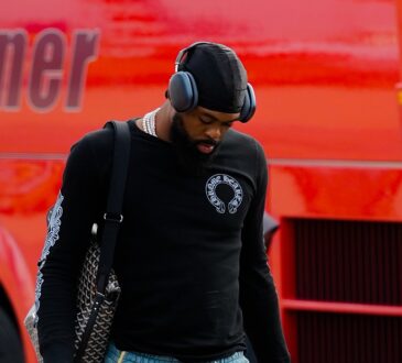 Dallas Mavericks forward Naji Marshall walks past a red bus on the tarmac, wearing headphones and carrying travel bags.