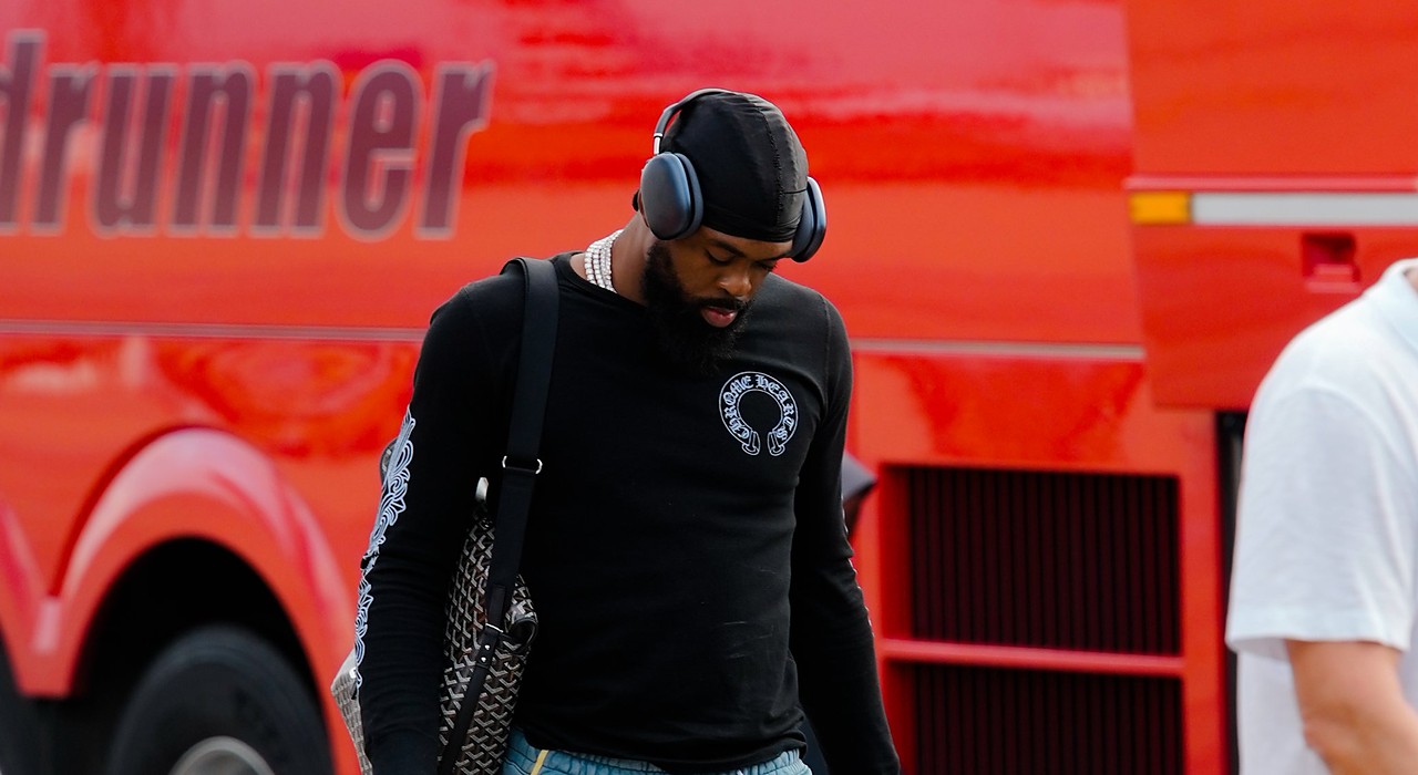Dallas Mavericks forward Naji Marshall walks past a red bus on the tarmac, wearing headphones and carrying travel bags.