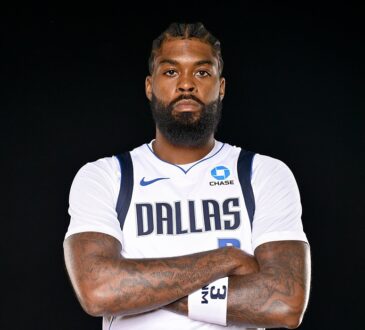 Mavericks forward Naji Marshall poses with his arms crossed in a white Dallas jersey against a black backdrop.