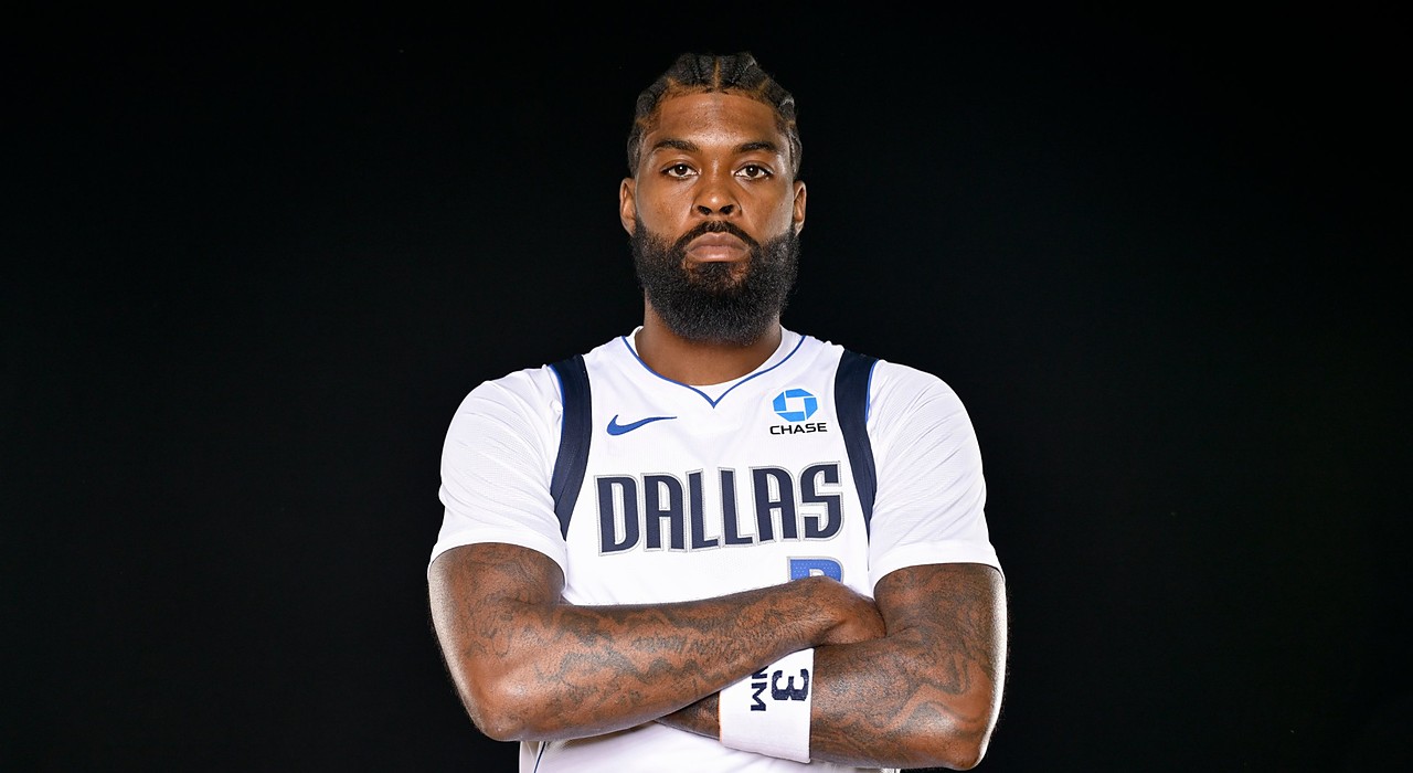 Mavericks forward Naji Marshall poses with his arms crossed in a white Dallas jersey against a black backdrop.