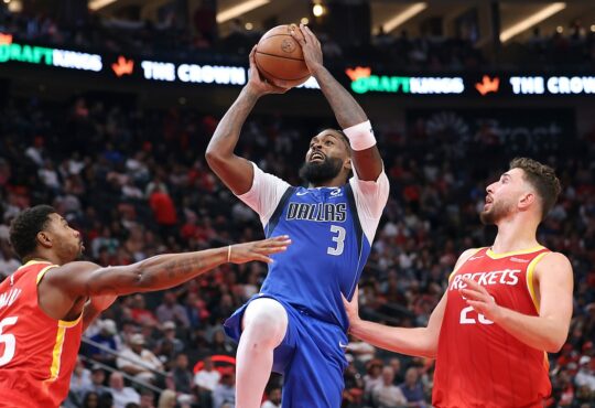 Dallas Mavericks forward Naji Marshall rises with the ball overhead between two Houston Rockets defenders.