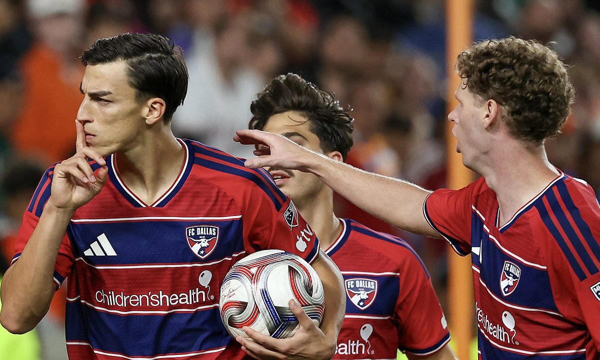 FC Dallas forward Petar Musa holds a finger to his lips while carrying the ball as teammates join him.