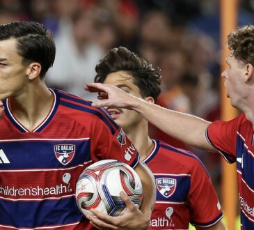 FC Dallas forward Petar Musa holds a finger to his lips while carrying the ball as teammates join him.