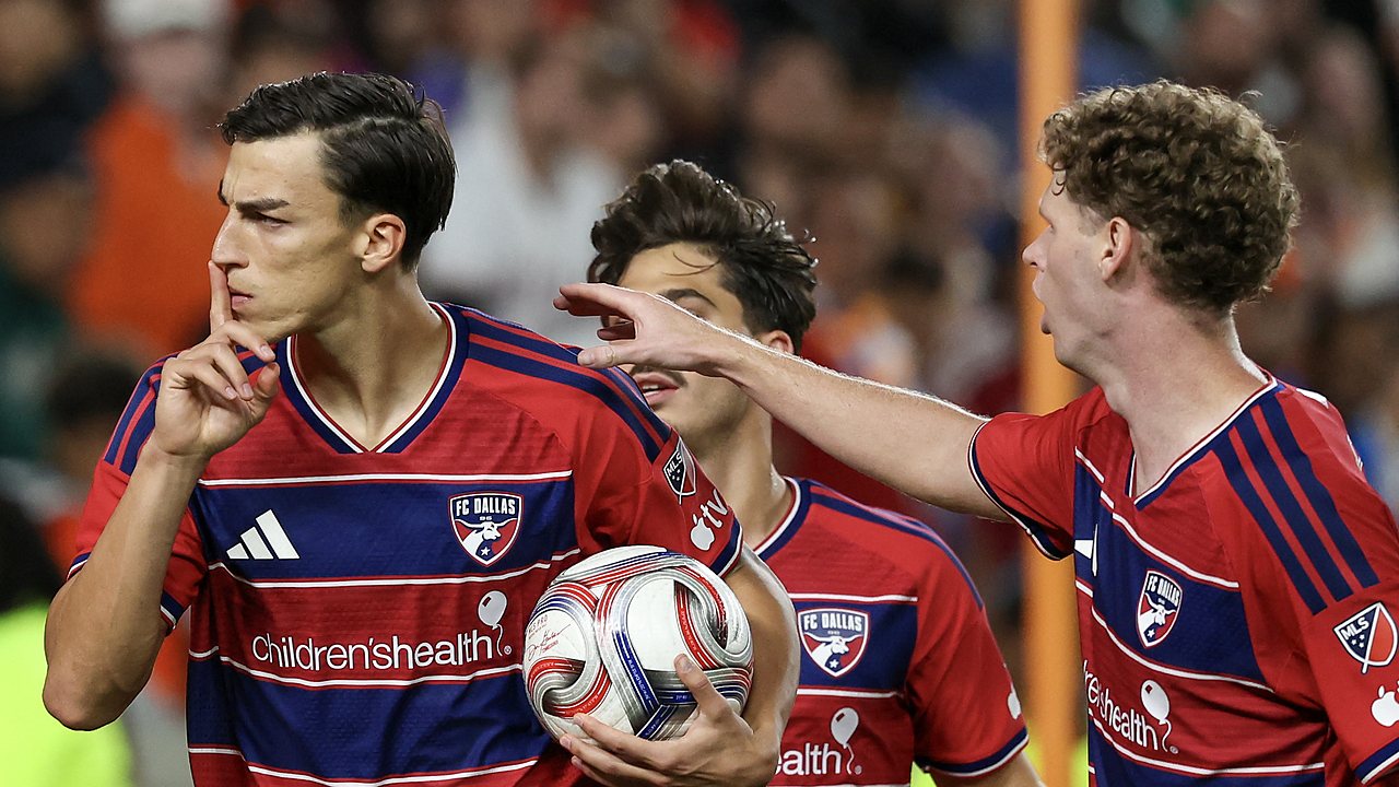 FC Dallas forward Petar Musa holds a finger to his lips while carrying the ball as teammates join him.