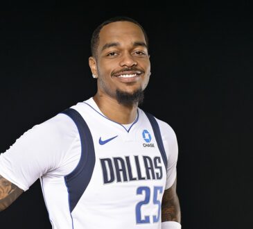 Dallas Mavericks forward P.J. Washington smiles with his hands on his hips in a white Dallas jersey against a black backdrop.