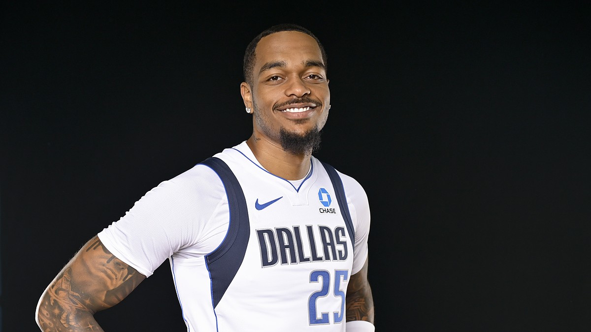 Dallas Mavericks forward P.J. Washington smiles with his hands on his hips in a white Dallas jersey against a black backdrop.