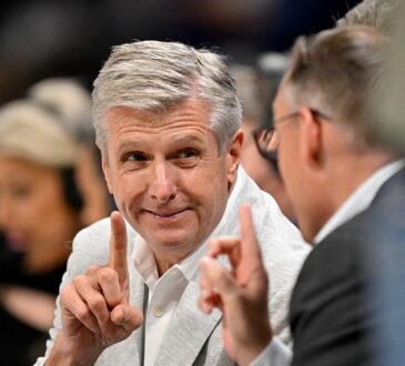 Dallas Mavericks CEO Rick Welts smiles and raises one finger as Dallas Wings CEO Greg Bibb, facing him, does the same.