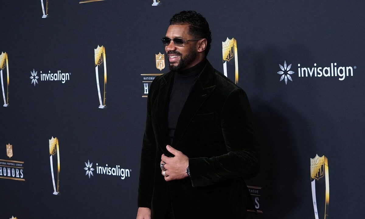 Former NFL quarterback Russell Wilson smiles on the NFL Honors red carpet in sunglasses and a black velvet suit.