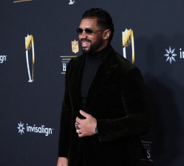 Former NFL quarterback Russell Wilson smiles on the NFL Honors red carpet in sunglasses and a black velvet suit.