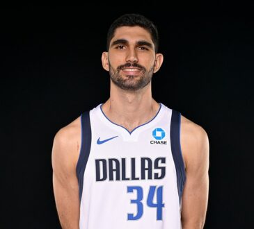 Dallas Mavericks forward-center Santi Aldama smiles with his hands behind his back in a white Dallas No. 34 jersey.