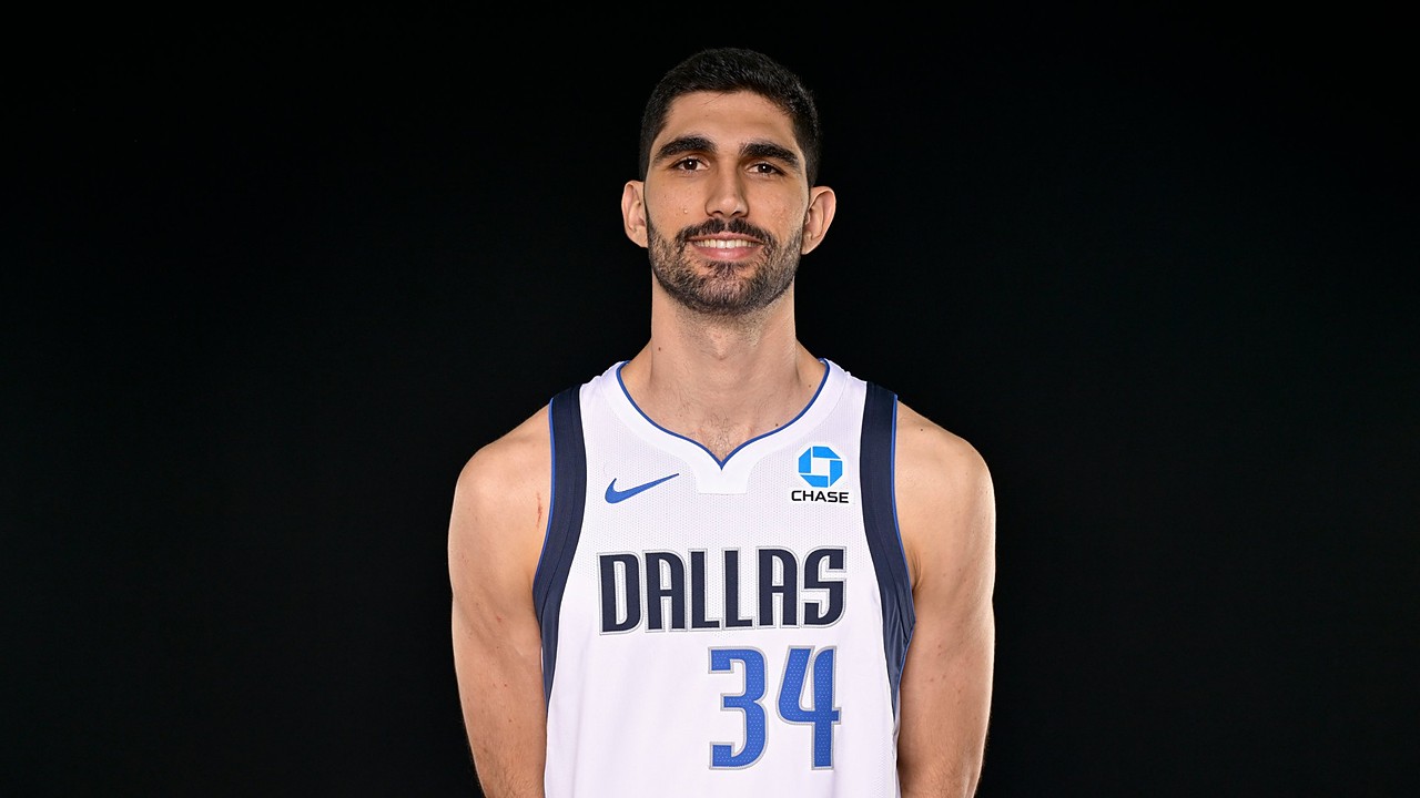 Dallas Mavericks forward-center Santi Aldama smiles with his hands behind his back in a white Dallas No. 34 jersey.