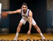 Dallas Mavericks forward-center Santi Aldama crouches in a wide dribbling stance with the ball on a hardwood court between studio lights.