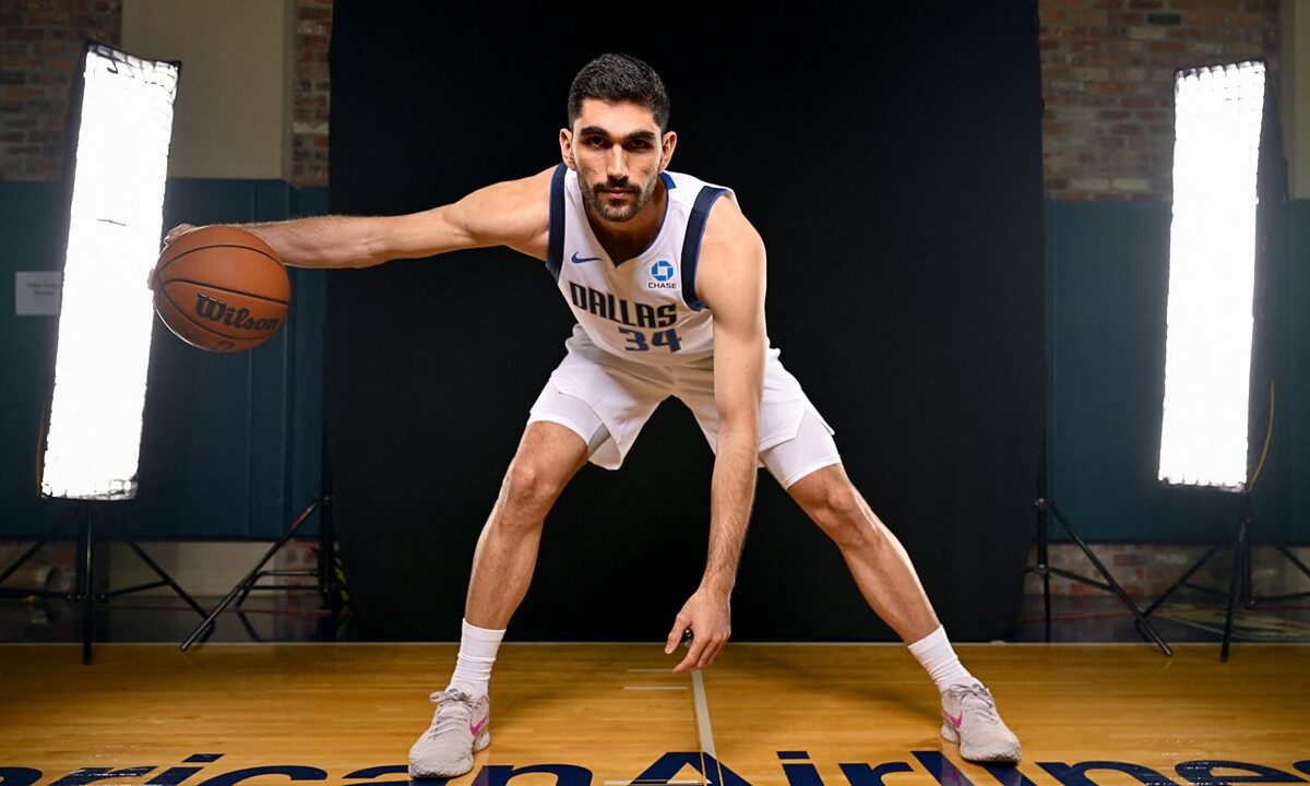 Dallas Mavericks forward-center Santi Aldama crouches in a wide dribbling stance with the ball on a hardwood court between studio lights.