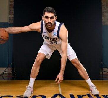 Dallas Mavericks forward-center Santi Aldama crouches in a wide dribbling stance with the ball on a hardwood court between studio lights.