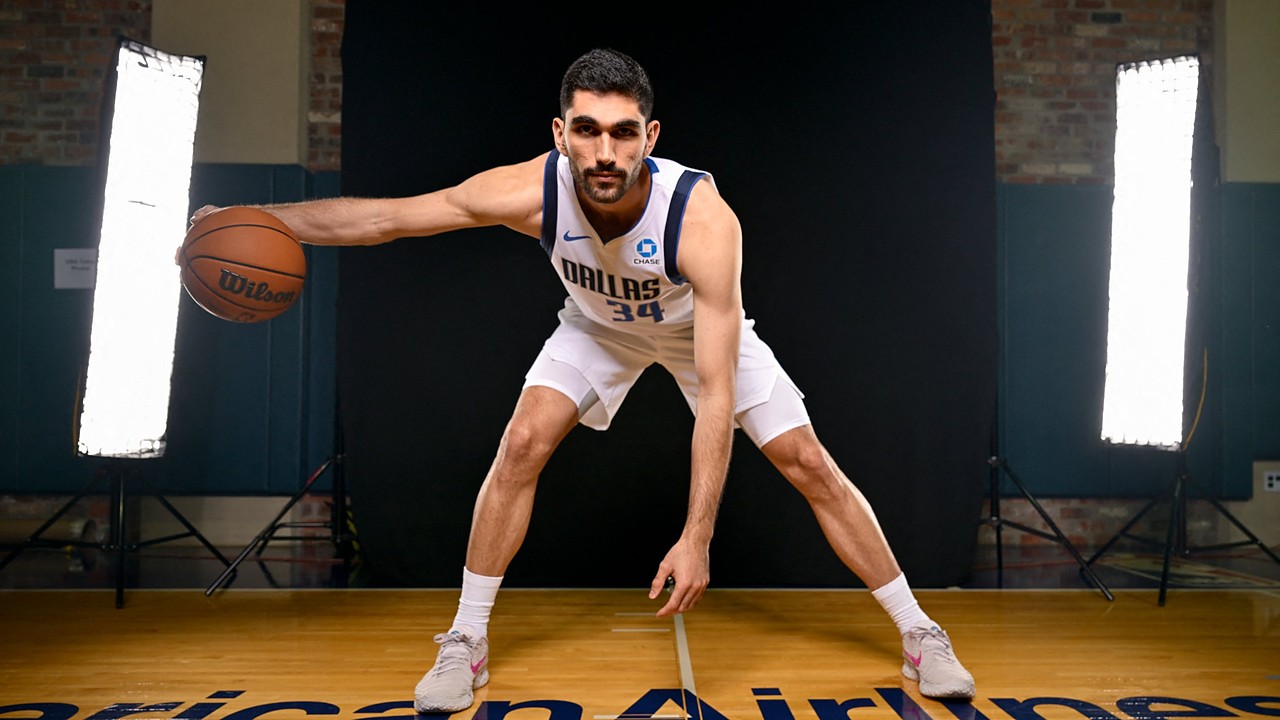 Dallas Mavericks forward-center Santi Aldama crouches in a wide dribbling stance with the ball on a hardwood court between studio lights.