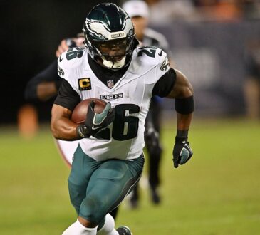 Philadelphia Eagles running back Saquon Barkley carries the ball upfield with a teammate and a defender trailing.