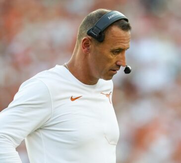 Texas head coach Steve Sarkisian looks down with a hand on his hip while wearing a headset on the sideline.
