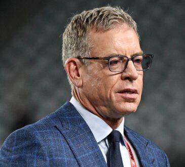 Former Dallas Cowboys quarterback Troy Aikman, in glasses and a blue plaid suit, looks on before the game.