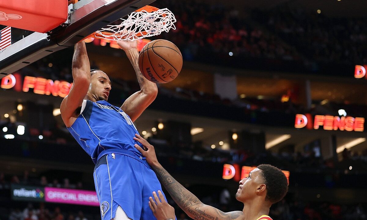 Dallas Mavericks forward Zaccharie Risacher hangs on the rim after a dunk as Houston Rockets forward Jabari Smith Jr. looks up.